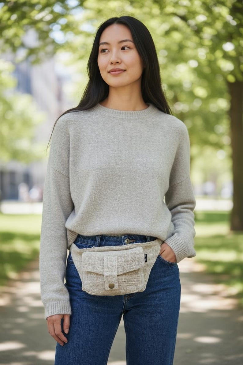 K&ouml;nnte beinhalten: Eine Frau tr&auml;gt einen hellgrauen Pullover und blaue Jeans mit einer beigen Bauchtasche. Die Bauchtasche hat eine rechteckige Form mit einer Klappe und einem Knopfverschluss. Der Hintergrund ist verschwommen mit B&auml;umen und einem Geb&auml;ude.