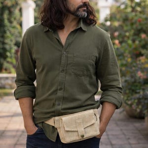 K&ouml;nnte beinhalten: Eine beigefarbene Bauchtasche mit Klappe und Knopfverschluss wird &uuml;ber einem gr&uuml;nen Button-Down-Hemd und dunkelblauen Jeans getragen. Die Bauchtasche ist aus einem gewebten Material gefertigt. Die Person steht drau&szlig;en in einem Garten.
