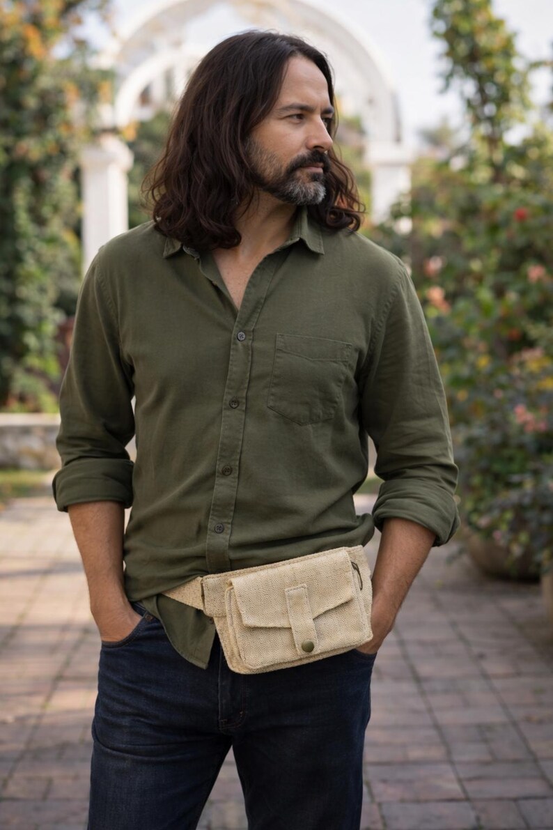 K&ouml;nnte beinhalten: Eine beigefarbene Bauchtasche mit Klappe und Knopfverschluss wird &uuml;ber einem gr&uuml;nen Button-Down-Hemd und dunkelblauen Jeans getragen. Die Bauchtasche ist aus einem gewebten Material gefertigt. Die Person steht drau&szlig;en in einem Garten.