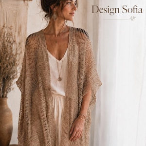 Könnte beinhalten: Eine Frau trägt einen beigefarbenen, offen gewebten Kimono-Cardigan über einem weißen Oberteil und einer Hose. Der Cardigan hat ein lockeres, fließendes Design. Der Text "Design Sofia" ist im oberen rechten Eck sichtbar. Der Hintergrund umfasst eine Vase und einen Makramee-Wandbehang.