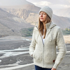May include: A woman wearing a light gray knit hat and a matching zip-up hooded sweater. The sweater has a textured pattern and a small brown patch on the lower right side. The background features a mountainous landscape and a riverbed.