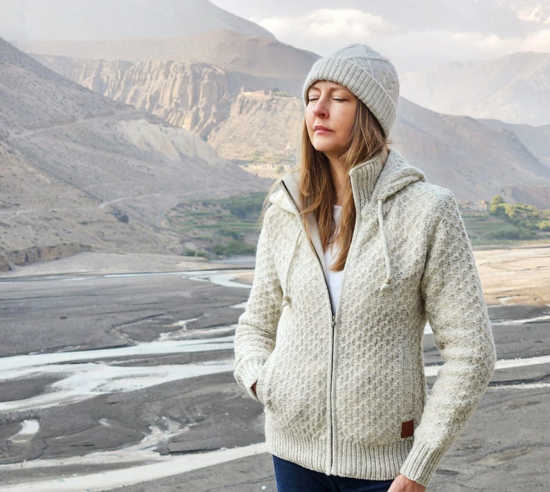 May include: A woman wearing a light gray knit hat and a matching zip-up hooded sweater. The sweater has a textured pattern and a small brown patch on the lower right side. The background features a mountainous landscape and a riverbed.