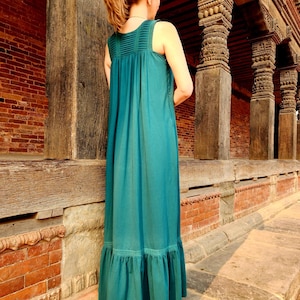 Pode incluir: Um vestido maxi turquesa com corpete plissado e bainha com babados. O vestido sem mangas &eacute; comprido e tem uma silhueta fluida. O vestido &eacute; usado ao ar livre em frente a um edif&iacute;cio de tijolo e madeira.