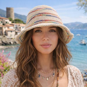 Peut inclure: Un chapeau seau en crochet beige avec des rayures multicolores. Le chapeau a un large bord. La personne sur la photo porte le chapeau et a de longs cheveux bruns ondulés. En arrière-plan, une scène côtière avec des bâtiments et la mer.