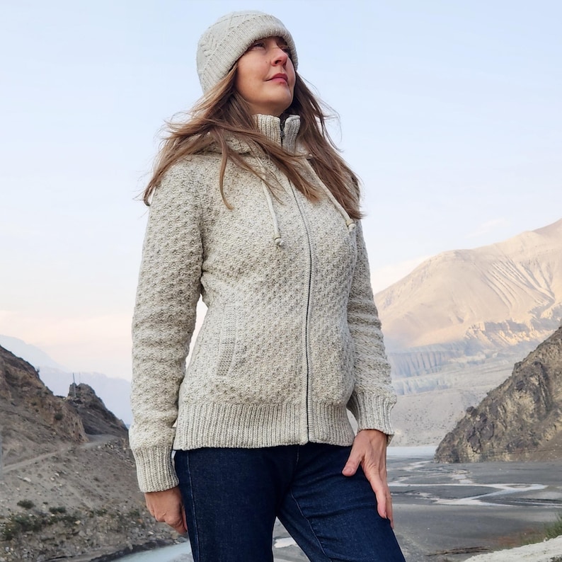 May include: A woman wearing a light gray knit zip-up hooded jacket and matching knit hat. The jacket has a textured pattern and a drawstring hood. She is also wearing dark blue jeans. The background features a mountainous landscape.