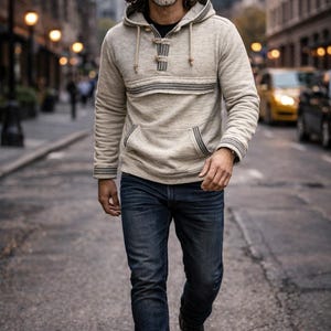 Op de afbeelding: Een persoon gekleed in een beige hoodie met een voorzak en een donkerblauwe jeans loopt door een stadsstraat. De hoodie heeft een decoratieve streep over de borst en een knoopsluiting. Hij draagt een zonnebril en zwarte laarzen.