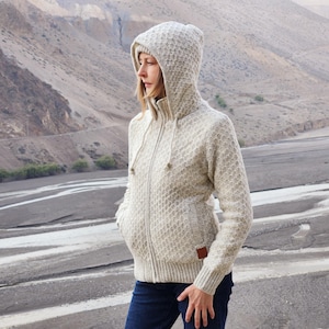 May include: A light beige hooded zip-up sweater with a textured pattern. The sweater has a drawstring hood, front pockets, and a small brown patch on the lower right side. The person is wearing dark blue jeans.