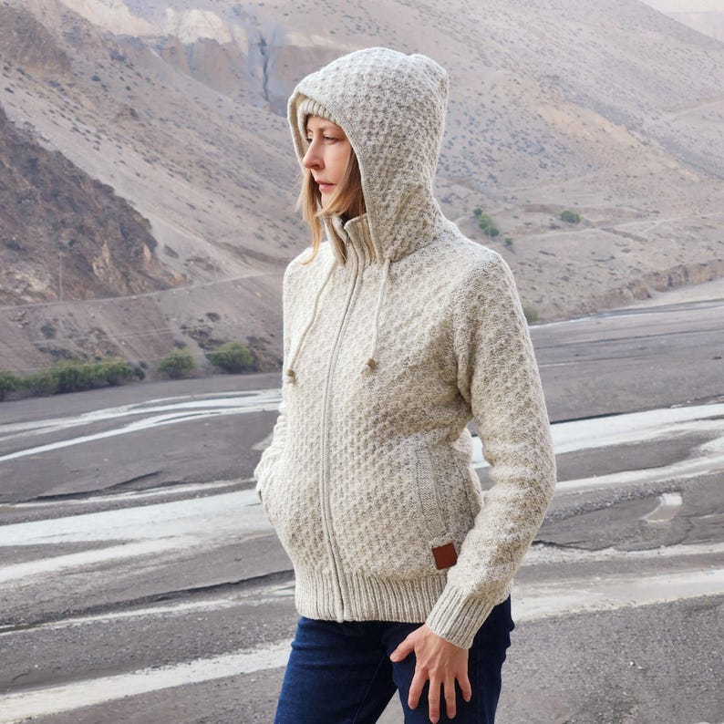 May include: A light beige hooded zip-up sweater with a textured pattern. The sweater has a drawstring hood, front pockets, and a small brown patch on the lower right side. The person is wearing dark blue jeans.