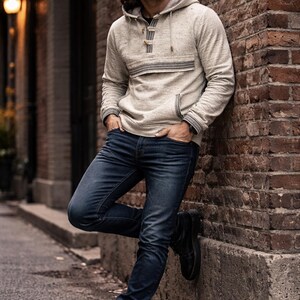 Op de afbeelding: Een lichtgrijze hoodie met een voorzak en een knoopsluiting. De hoodie wordt gecombineerd met een donkerblauwe jeans en zwarte laarzen. De persoon leunt tegen een bakstenen muur.