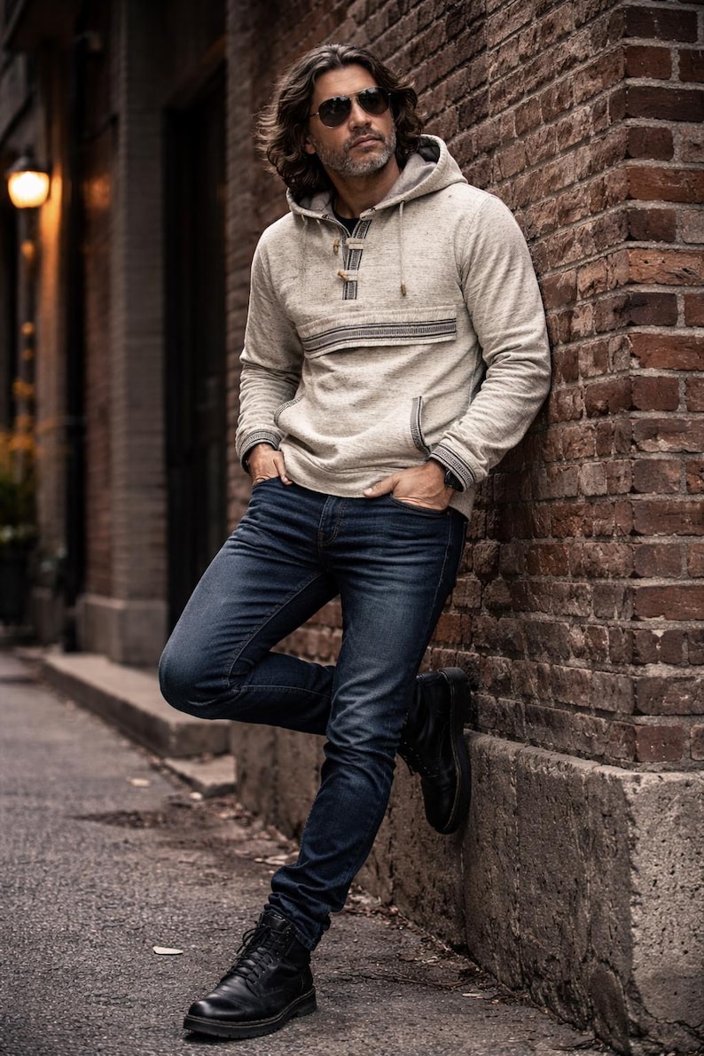 Op de afbeelding: Een lichtgrijze hoodie met een voorzak en een knoopsluiting. De hoodie wordt gecombineerd met een donkerblauwe jeans en zwarte laarzen. De persoon leunt tegen een bakstenen muur.