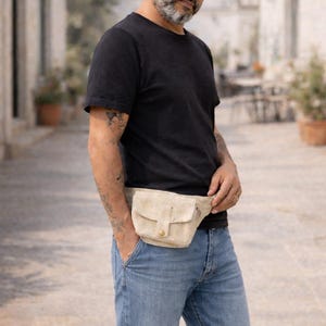 K&ouml;nnte beinhalten: Eine beigefarbene Bauchtasche mit Klappe und Knopfverschluss. Die Tasche wird um die Taille &uuml;ber einem schwarzen T-Shirt und einer blauen Jeans getragen. Die Bauchtasche hat eine kleine Tasche auf der Vorderseite.
