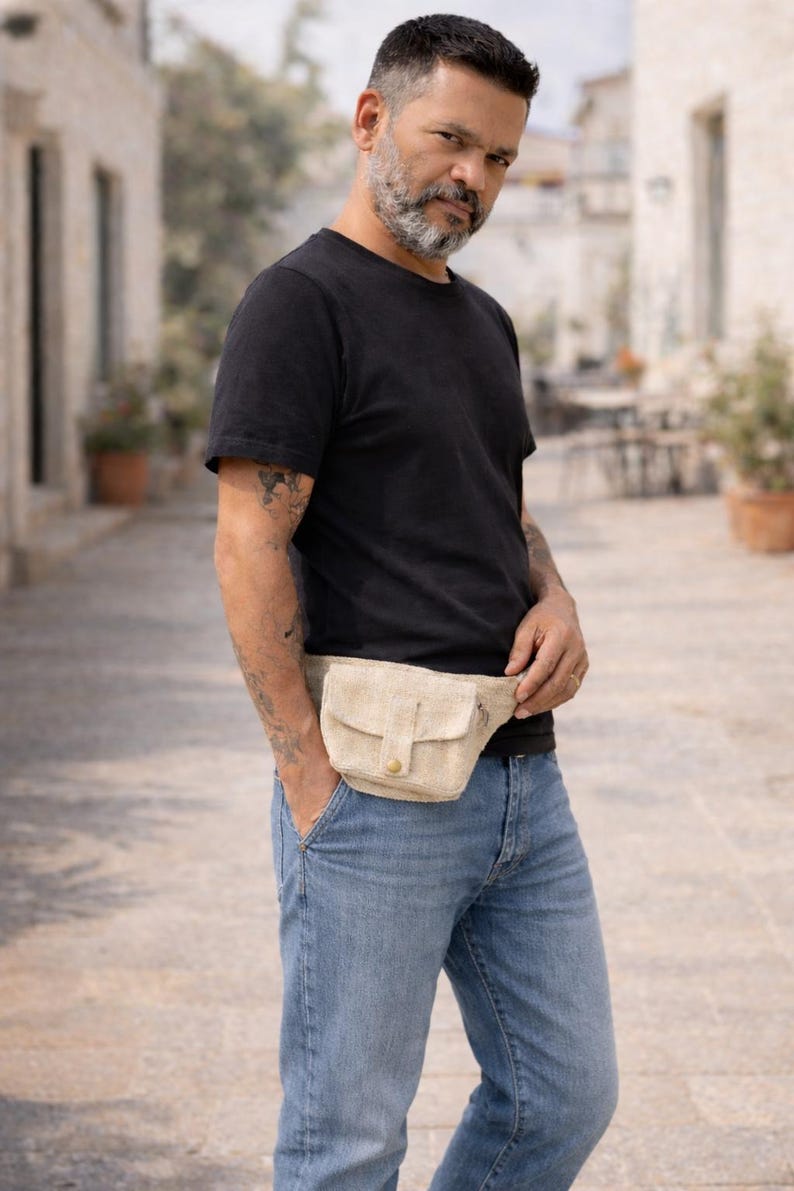K&ouml;nnte beinhalten: Eine beigefarbene Bauchtasche mit Klappe und Knopfverschluss. Die Tasche wird um die Taille &uuml;ber einem schwarzen T-Shirt und einer blauen Jeans getragen. Die Bauchtasche hat eine kleine Tasche auf der Vorderseite.