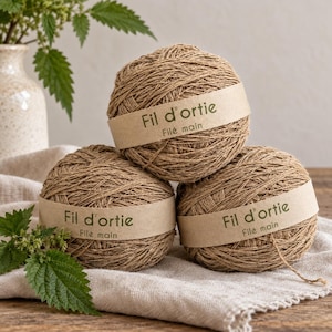 Pode incluir: Três novelos redondos de fio castanho-claro e texturizado empilhados. Cada novelo apresenta uma faixa de papel bege com o texto em verde escuro Fil d'ortie e Filé main. O fio tem uma aparência natural e fibrosa com fios visíveis.