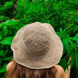 May include: A woven, beige sun hat with a wide brim. The hat is made of a natural fiber and has a circular pattern on the crown. The hat is worn outdoors, surrounded by green foliage.