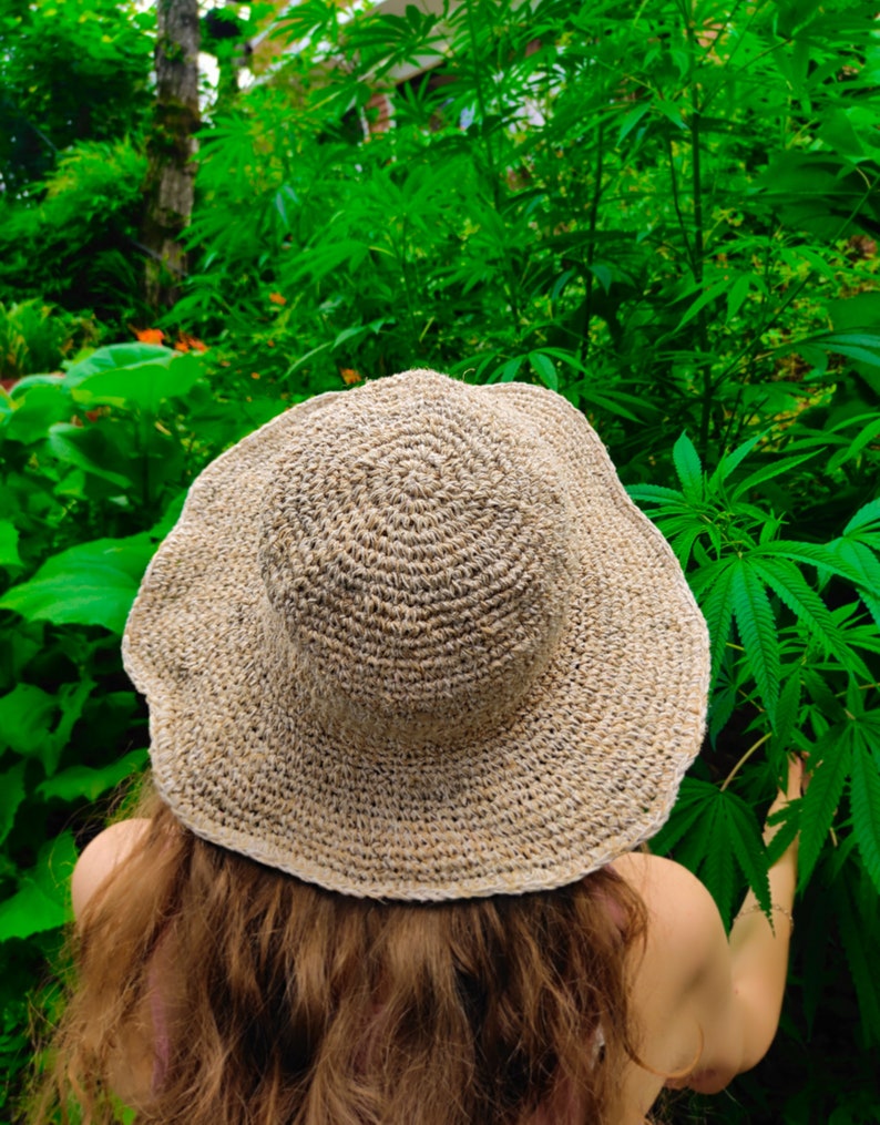 May include: A woven, beige sun hat with a wide brim. The hat is made of a natural fiber and has a circular pattern on the crown. The hat is worn outdoors, surrounded by green foliage.