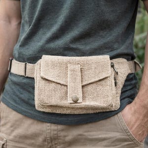 K&ouml;nnte beinhalten: Eine beigefarbene H&uuml;fttasche aus Hanf mit Klappenverschluss und einer kleinen Rei&szlig;verschlusstasche. Der verstellbare G&uuml;rtel ist ebenfalls beige. Die Tasche wird um die Taille &uuml;ber einem dunkelgr&uuml;nen Hemd und einer khakifarbenen Hose getragen.