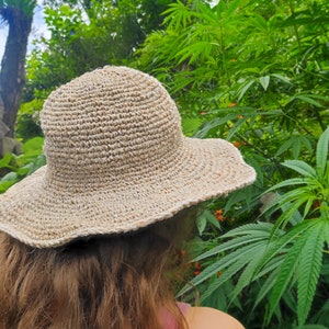 May include: A beige, woven sun hat with a wide brim. The hat is made of a natural fiber and has a slightly wavy edge. The background features lush green foliage and orange flowers.