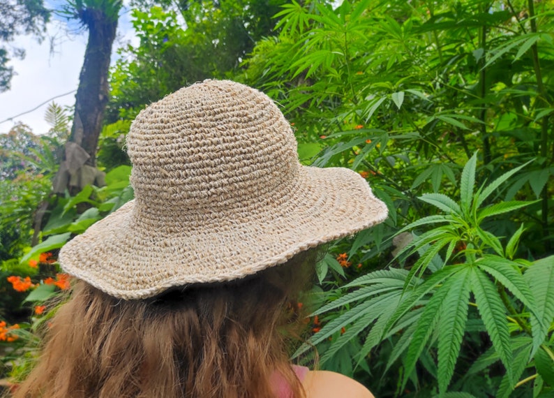 May include: A beige, woven sun hat with a wide brim. The hat is made of a natural fiber and has a slightly wavy edge. The background features lush green foliage and orange flowers.