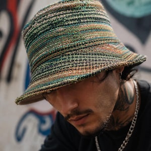 May include: A bucket hat featuring a multi-colored woven texture with horizontal stripes in shades of green, orange, brown, and black. The hat has a structured crown and a downward-sloping brim, crafted from a thick, textured fabric material.
