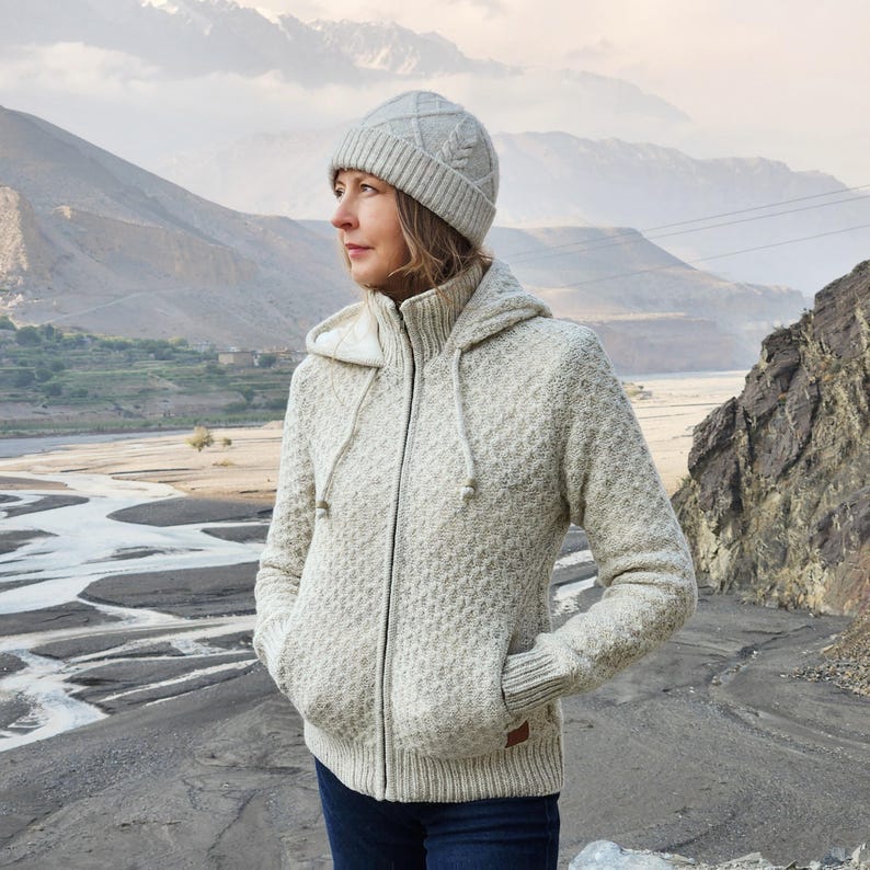 May include: A light gray hooded zip-up sweater and matching knit cap. The sweater has a textured pattern, a drawstring hood, and front pockets. The cap features a cable knit design. The outfit is suitable for outdoor activities.