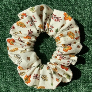 May include: A white fabric scrunchie with a fall-themed print of orange, yellow, and green leaves and red berries.