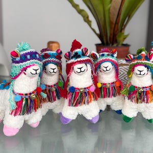 May include: A group of white plush alpaca dolls, each adorned with a colorful knitted hat and matching tassel accents. The dolls have small bells around their necks and are arranged on a reflective surface.