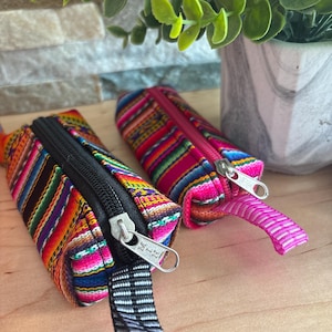 Può includere: Due piccole pochette rettangolari con esterni in tessuto intrecciato colorato. Una ha una cerniera e un tirante neri, l'altra una cerniera e un tirante rosa. Le pochette sono decorate con un motivo a righe in vari colori.