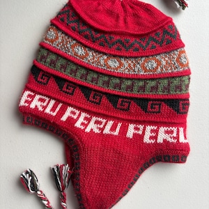 May include: A red knitted chullo hat with earflaps and a braided tassel at the crown. The design incorporates horizontal bands of geometric patterns in green, black, orange, and white, with the word PERU repeated in white text across the centre.