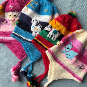 May include: Four knitted earflap hats with pom-poms and embroidered animal appliqués. The hats feature horizontal stripes in pink, blue, red, yellow, and white. Each hat displays a unique stitched character, including a llama, a goat, and a rabbit.