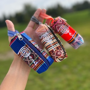 May include: Three small cylindrical zippered pouches featuring woven textile patterns in blue, orange, and red. Each pouch displays the word Peru embroidered in white script with a spiral design. Each pouch includes a small fabric wrist strap.
