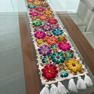 Könnte beinhalten: Weißer, bestickter Tischläufer mit einem lebendigen Blumenmuster. Der Läufer zeigt bunte Blumen in Rot-, Rosa-, Orange-, Gelb-, Grün-, Blau- und Lilatönen. Weiße Quasten zieren jedes Ende.