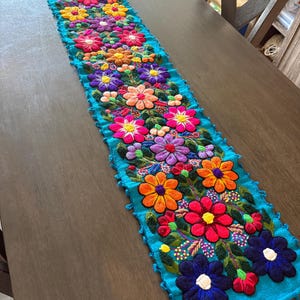 May include: A long, rectangular turquoise table runner featuring dense, multi-colored floral embroidery. The design includes raised, vibrant flowers in shades of pink, orange, purple, yellow, and red, accented with green leaves and small tassels.