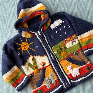 May include: A navy blue knit zip-up hoodie featuring a colorful landscape scene with a yellow sun, a white cloud with rain, a small house, a white llama, and a gray turtle. The sleeves and hood are accented with horizontal stripes of yellow, green, and red.