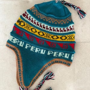 Puede incluir: Un gorro de punto color verde azulado con orejeras que presenta un patrón repetitivo de llamas, formas geométricas y la palabra PERU impresa en verde claro. El gorro incluye una borla trenzada en la parte superior y dos borlas trenzadas en los extremos de las orejeras, en rojo, blanco y negro.