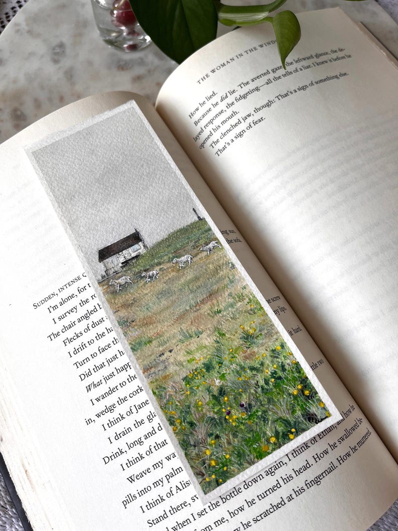 May include: A hand-painted bookmark depicting a rural scene with a white cottage, sheep, and wildflowers under a cloudy sky. The bookmark is set within an open book. A unique gift for book lovers. The bookmark is approximately 15 cm in length.