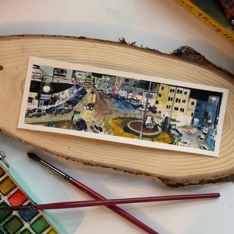 May include: Watercolour painting of a city street at night. The artwork shows buildings, cars, and streetlights in a long, rectangular format. The painting is on white paper, set on a wooden surface, with paintbrushes and a watercolour palette in the foreground.