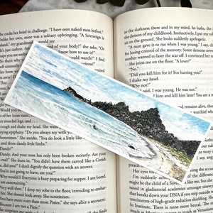 May include: A rectangular bookmark with a watercolour painting of a coastal scene, showing a rocky shoreline, ocean waves, and a blue sky. The bookmark is on an open book.