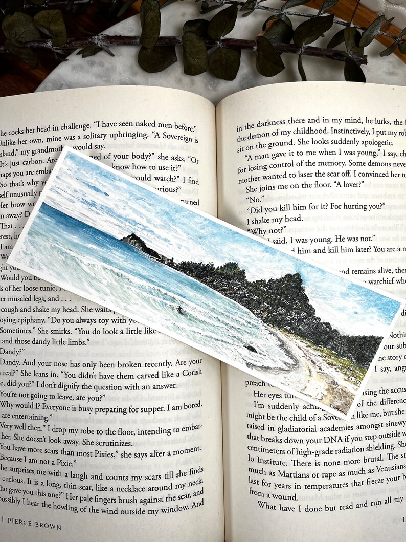 May include: A rectangular bookmark with a watercolour painting of a coastal scene, showing a rocky shoreline, ocean waves, and a blue sky. The bookmark is on an open book.