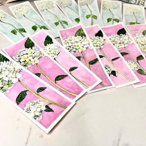 May include: Watercolor bookmarks depicting white hydrangea flowers with green leaves and stems. The bookmarks have a pink or blue watercolour background. Books are visible in the background.