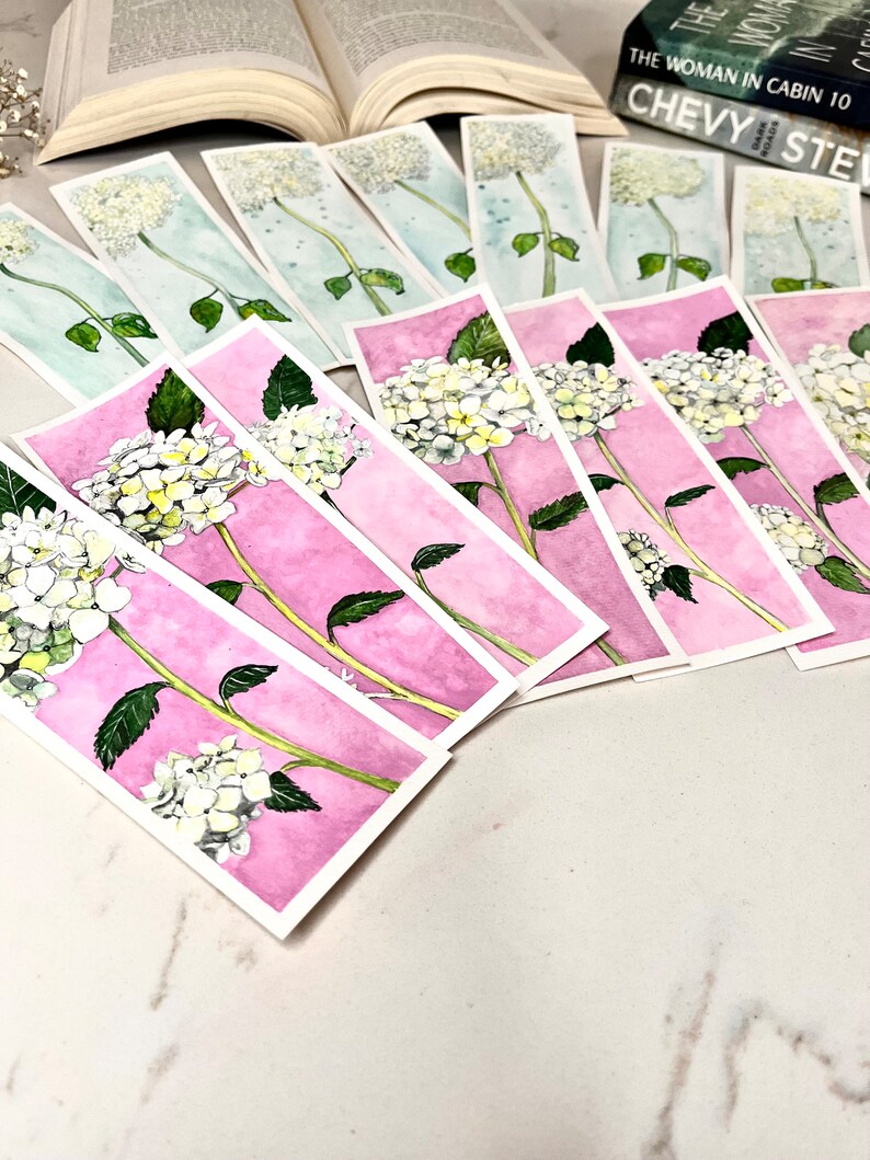 May include: Watercolor bookmarks depicting white hydrangea flowers with green leaves and stems. The bookmarks have a pink or blue watercolour background. Books are visible in the background.