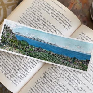 May include: A rectangular watercolour bookmark depicting a landscape with a lake, trees, and mountains. The bookmark is placed on open book pages, with text visible. The scene evokes a sense of calm.