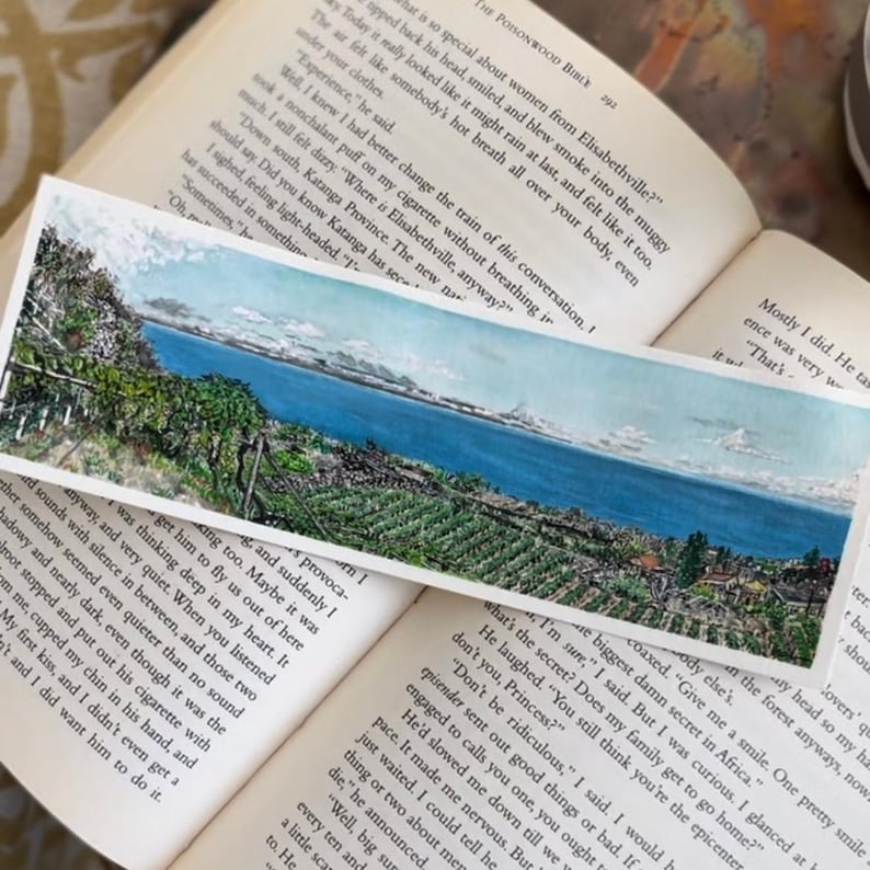 May include: A rectangular watercolour bookmark depicting a landscape with a lake, trees, and mountains. The bookmark is placed on open book pages, with text visible. The scene evokes a sense of calm.