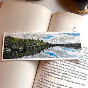May include: A rectangular watercolour bookmark depicting a tranquil river scene with trees and a cloudy sky. The bookmark lies on an open book, with the number 29 visible. The image suggests a peaceful, natural setting.