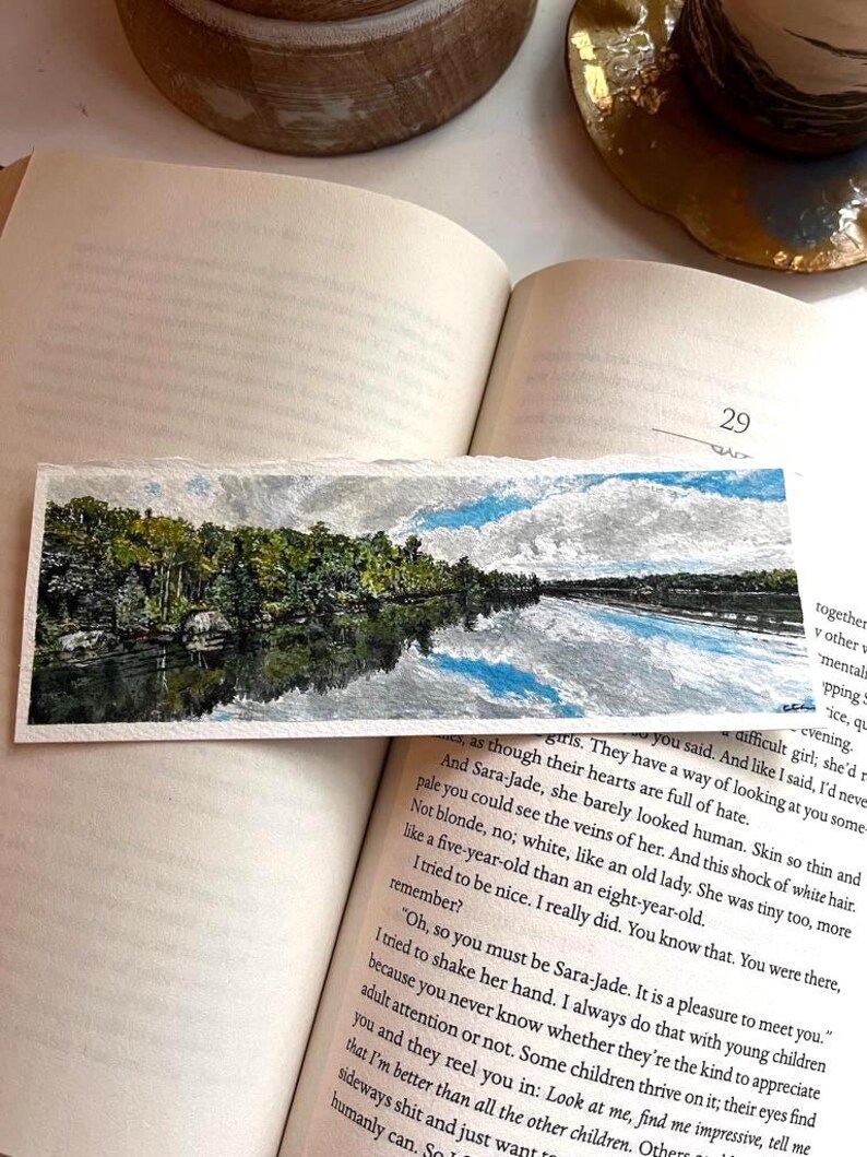 May include: A rectangular watercolour bookmark depicting a tranquil river scene with trees and a cloudy sky. The bookmark lies on an open book, with the number 29 visible. The image suggests a peaceful, natural setting.