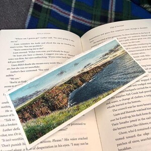 May include: A bookmark depicting a coastal scene with cliffs, sea, and sky. The bookmark is on an open book with text. The image has a watercolour painting style.