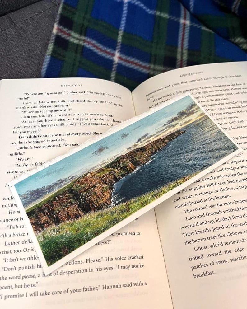 May include: A bookmark depicting a coastal scene with cliffs, sea, and sky. The bookmark is on an open book with text. The image has a watercolour painting style.