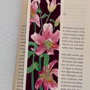 May include: A bookmark with a floral design of pink lilies with green stems and leaves on a dark purple background. The bookmark is rectangular and is on an open book.