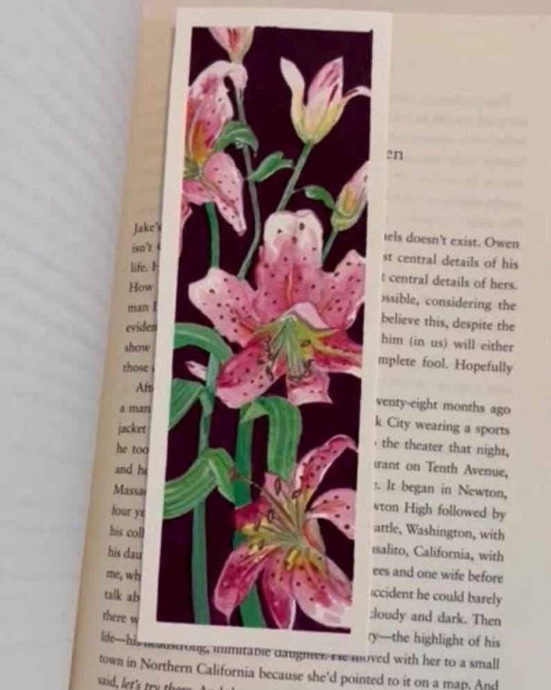 May include: A bookmark with a floral design of pink lilies with green stems and leaves on a dark purple background. The bookmark is rectangular and is on an open book.