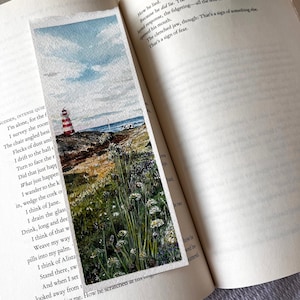 May include: A watercolour bookmark depicting a coastal landscape with a red and white striped lighthouse, blue sky, and sea. The bookmark is rectangular and shows a detailed painting. The bookmark is placed in an open book.