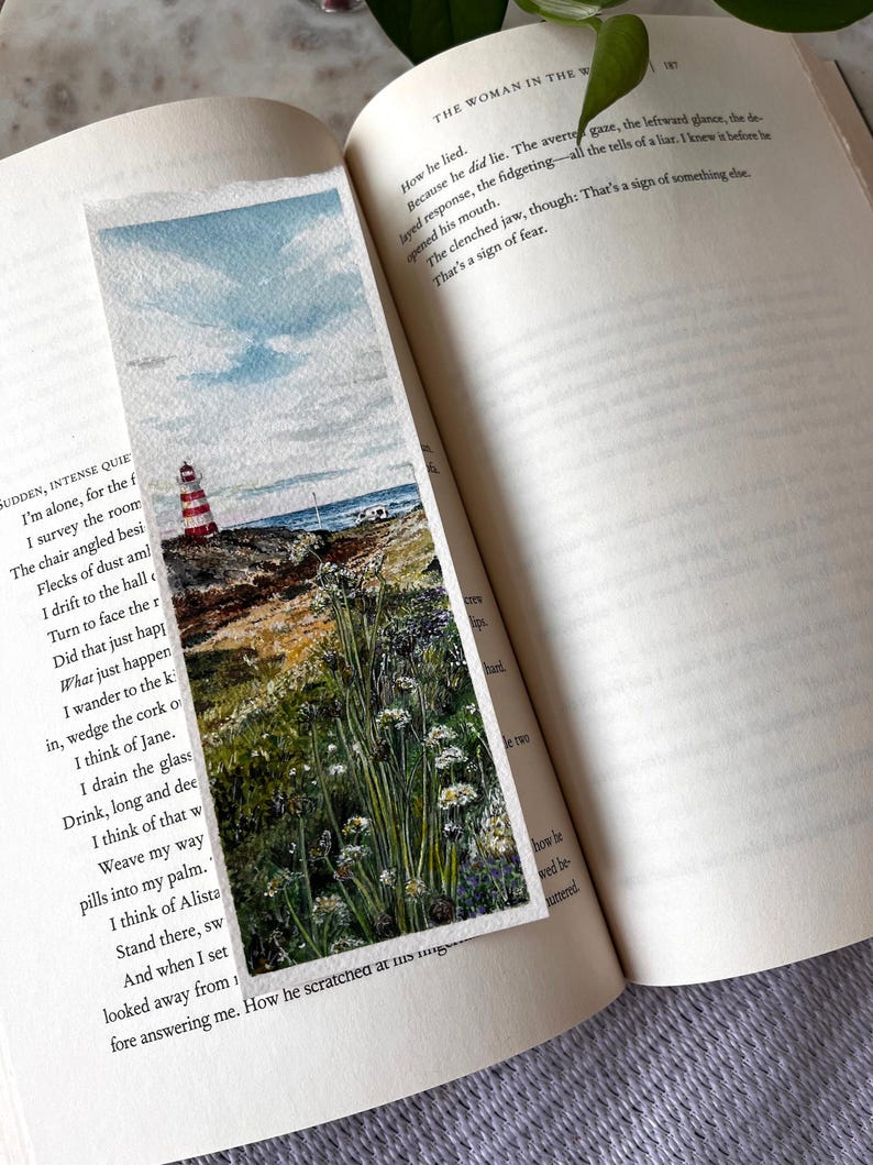 May include: A watercolour bookmark depicting a coastal landscape with a red and white striped lighthouse, blue sky, and sea. The bookmark is rectangular and shows a detailed painting. The bookmark is placed in an open book.